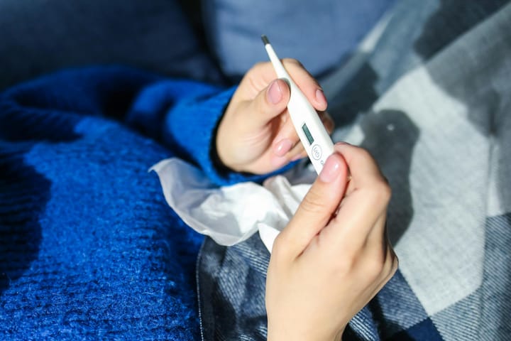 Someone lays in bed with a tissue and looking at a small thermometer
